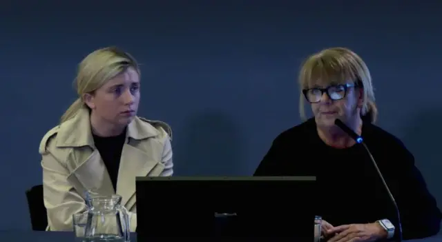 A woman with blonde hair, wearing a black top and cream coloured coat. She is sitting to the left of a woman with blonde hair, wearing a black top.