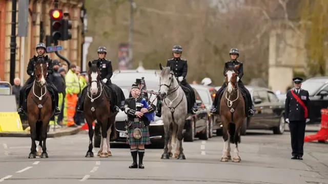 A man in a kilt plays the bagpipes in front of a procession of four horses. Behind them are several funeral cars.