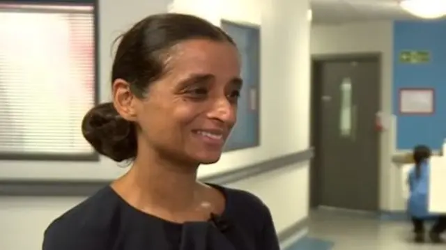 Standing in a hospital corridor, Gillmore smiles. She has brown hair in a low bun and wears a black top