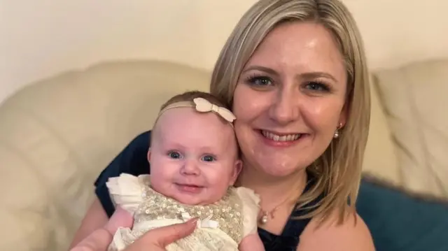 A woman smiling at the camera as she holds her baby who is also smiling