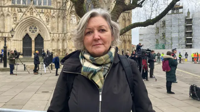 A woman with short grey hair, wearing a black jacket and green scarf, stands outside York Minster