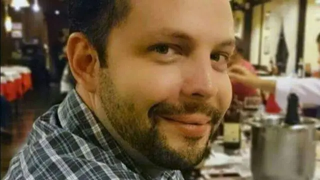 Headshot of Jairo smiling at the camera. He wears a plaid shirt and sits in a restaurant