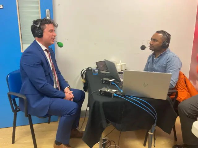 Two men sit in seats in a room with a blue door. On the left, Landstrom sits in a blue suit with headphones over his head which have a green mic attached. At a desk opposite sits another man in a blue denim shirt, with two laptops open in front of him. He also wears headphones with a microphone