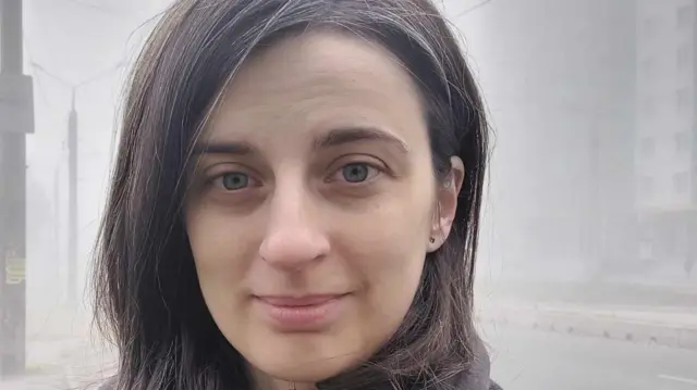 A woman smiles in front of a foggy background where you can see a road and a building.