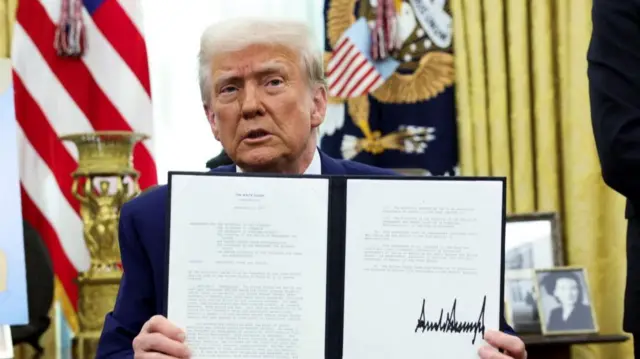 Trump sits at a desk holding a signed executive order