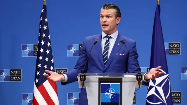 Pete Hegseth, wearing a blue suit, speaks behind a podium with the flags of Nato and the USA on either side.