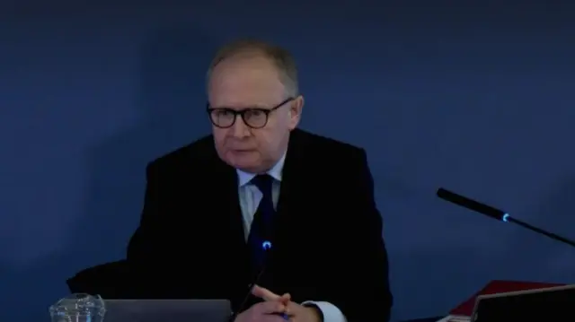 A man wearing glasses, a blue shirt, tie and black jacket sitting at a desk