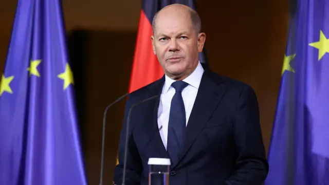 German Chancellor Olaf Scholz stands in front of the blue European union flag.