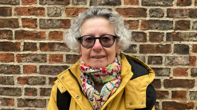 An older woman in a yellow jacket and colourful scarf stands against a brick wall.