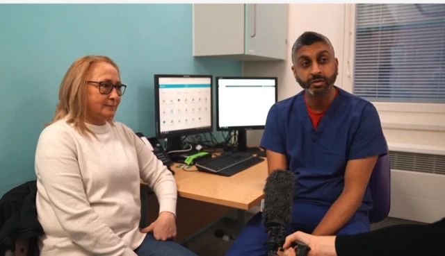 Emily sits on the left wearing a white jumper, she has short blonde hair and black glasses. On her left is the doctor, wearing blue scrubs. He has facial hair and sits at a desk with two monitors on it. A press microphone is being held to him as he speaks