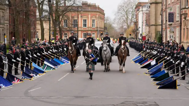 A man in a kilt plays the bagpipes in front of a procession of four horses. Police officers line either side of the street and point their flags to the ground.