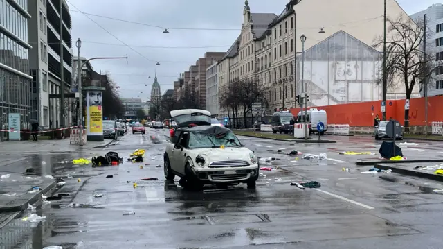 A white mini cooper sits in the middle of a roadway. It is heavily damaged and has a broken headlamp, a dented roof and its front bumper looks twisted. Debris - including several high-vis vests - is strewn across the roadway.