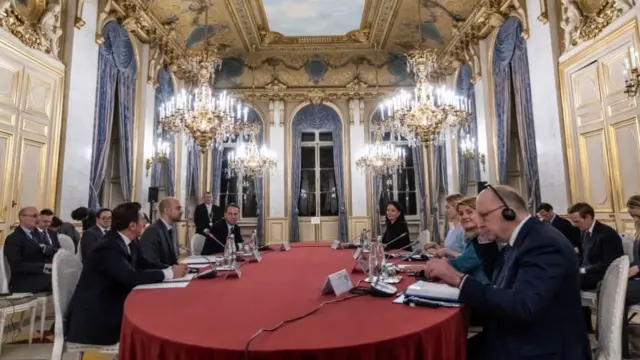 European defence leaders sitting down at table covered by red cloth attend meeting in Paris. They're in a ornate room with crystal and gold candelabra