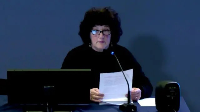 A woman with black curly hair, wearing glasses and a black top. She is holding a piece of paper and is sitting at a desk