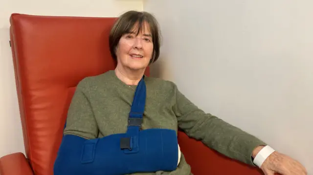 Patricia Nolan, 82, wearing a green jumper sits on an orange leather armchair, resting her right arm in a blue sling