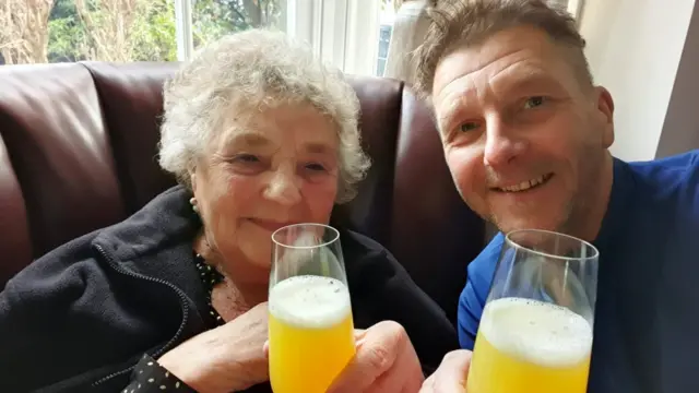 An older lady with curly grey hair next to a middle aged man. They both smile and hold prosecco glasses with orange liquid to the camera