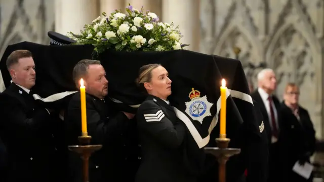 Pallbearers carry the coffin, draped in a police flag, into the Minster, which is lit with candles.