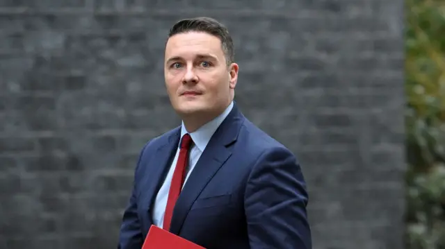 Wes Streeting, wearing a blue suit and holding a red folder, looks to the right past the camera