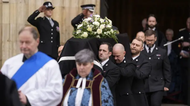 Pallbearers carry out coffin, draped in black and white flowers