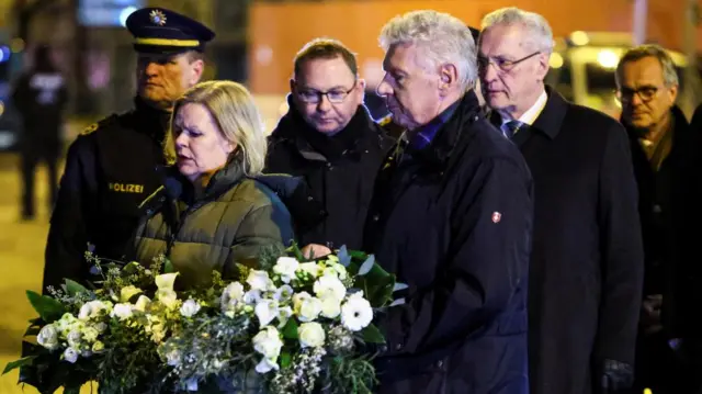 German Interior Minister Nancy Faeser, Munich mayor Dieter Reiter, Bavarian interior Minister Joachim Herrmann and public sector workers union Verdi's leader Frank Werneke visit the scene. it is night time and faeser is carrying a large bouquet of white flowers.