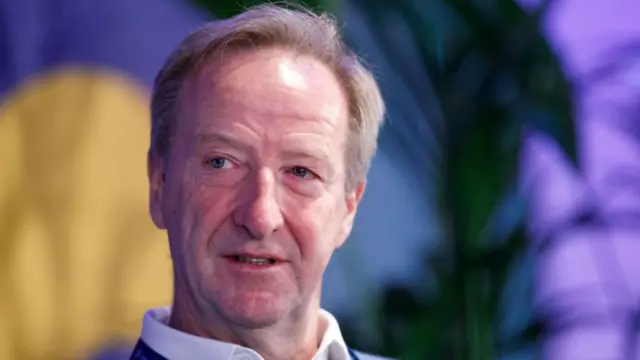 Former MI6 chief Sir Alex Younger close up as he sits down. A plant is visible to his left in the blurred background