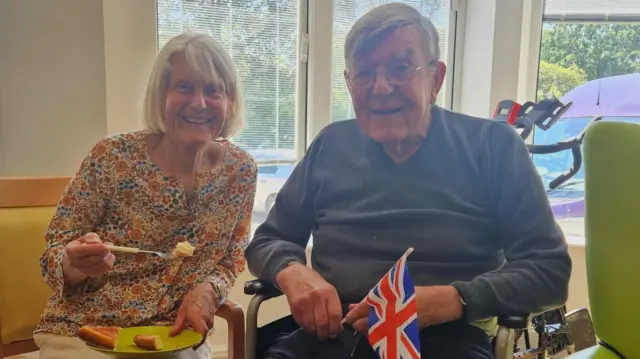 Woman in a floral top eating some cake next to a man in a blue jumper holding a small Union Jack flag.