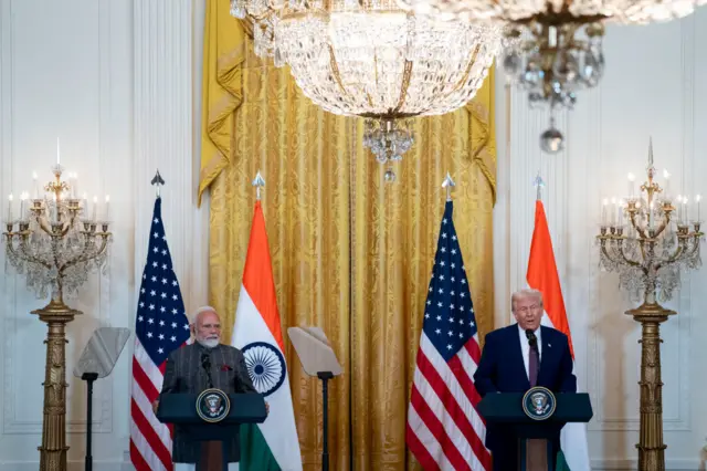Two men stand at podiums in front of gold curtain