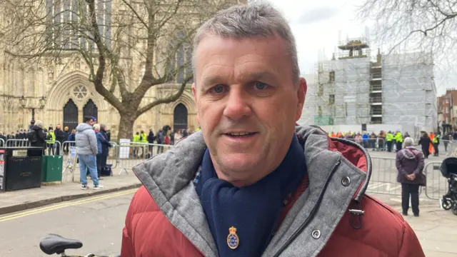 Man with short grey hair, in a red jacket and blue scarf with police badge on, stands outside the barriers at York Minster.