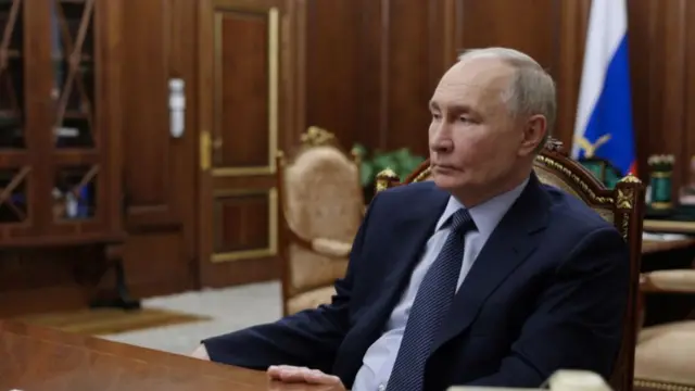 Vladimir Putin sitting down at wooden table in dark blue suit, blue shirt and polka-dots tie, his left hand grasping the table in front of him