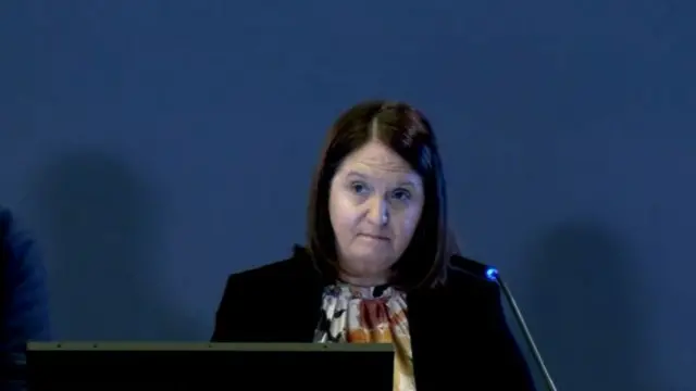 A woman with brown hair wearing a blouse and a black jacket. She is sitting at a desk.