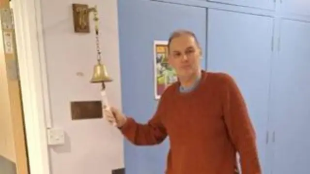 Man in red jumper ringing a bell attached to a hospital-ward wall, there are blue cupboards behind him