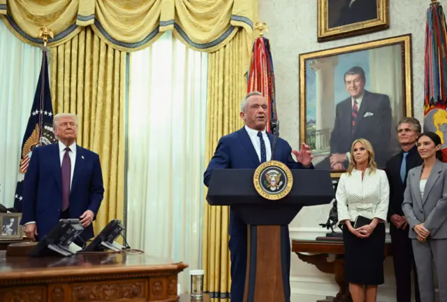Robert F Kennedy Jr spekaing inside the Oval office of the White House after being sworn in. President Donald Trump is standing nearby him at a desk, as is RFK's wife Cheryl Hines is behind to the right