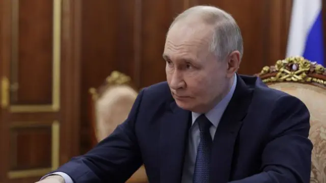 Vladimir Putin mid-shot as he sits down on brocade and gold-decorated wooden chair, his arms extended as they rest of a table. He's wearing a dark blue suit, blue shirt and blue tie