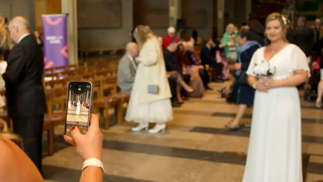 A woman in a white dress out of focus while a friend takes a picture on her phone