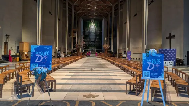 The inside of Coventry Cathedral with dozens of wooden chairs lined up, facing each other and two easels at the front with "We Still Do" posters on them.