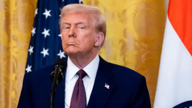 Donald Trump looks sideways to his left as he waits on stage, a yellow brocade curtain and US flag behind him