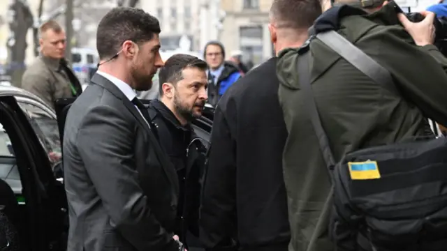 Zelensky exits car as he arrives in Munich. He's wearing total black and is surrounded by two men in suit with singular headphones, a single photographer with a Ukrainian patch on the bag to his side