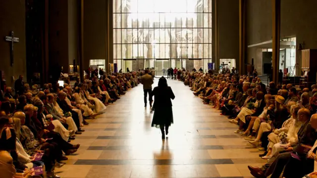 A brightly lit cathedral room with couple lines up in chairs either side of an aisle with sun shining through a large window at the end