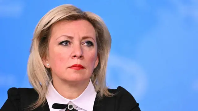 Maria Zakharova sitting down as she listens during her daily press briefing. She's wearing a black blazer, white shirt with a thin black bow around the neck