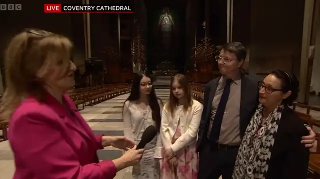 A BBC reporter in a pink suit with a couple and their children inside a large stone cathedral building with wooden chairs lined behind them