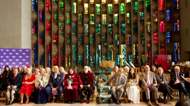 A row of couples sat in front of a number of multi-coloured windows at Coventry Cathedral