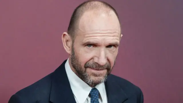 Ralph Fiennes posing for a photo, wearing a navy suit and tie and white shirt, against a purple wall