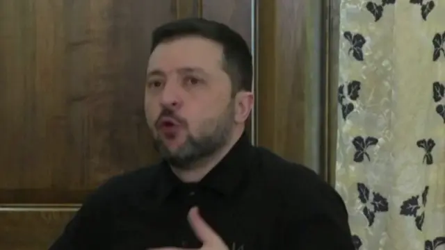 Zelensky sitting down in front of wooden door and curtain as he addresses reporters from inside a hotel in Munich