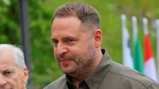 Andriy Yermak close up as he walks outdoors wearing a dark military green shirt, portion of a man's head visible to his right and blurred flags behind him