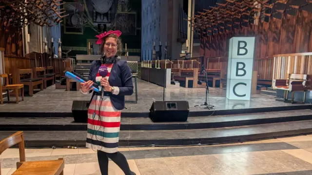 Reporter Rosie Eaton wearing a pink fascinator, a striped dress and holding a purple microphone inside the main room in Coventry Cathedral