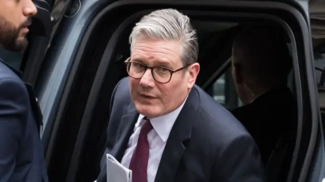 Keir Starmer looks straight ahead as he steps out of a black car wearing a black suit, white shirt and burgundy patterned tie