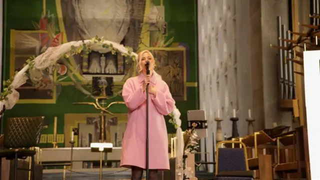 Pixie Lott wearing a pink dress sings into a black microphone in the cathedral. She has blonde hair