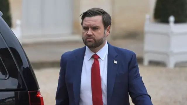 JD Vance walking around a black car as he arrives at Elysee Palace in Paris wearing a blue suit, white shirt