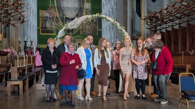 A large group of people, all stood in front of a flora arch on a stage at Coventry Cathedral