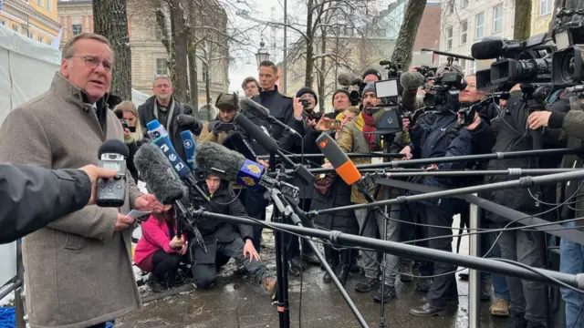 German Defence Minister Boris Pistorius, wearing a brown coat, speaks into a cluster of microphones